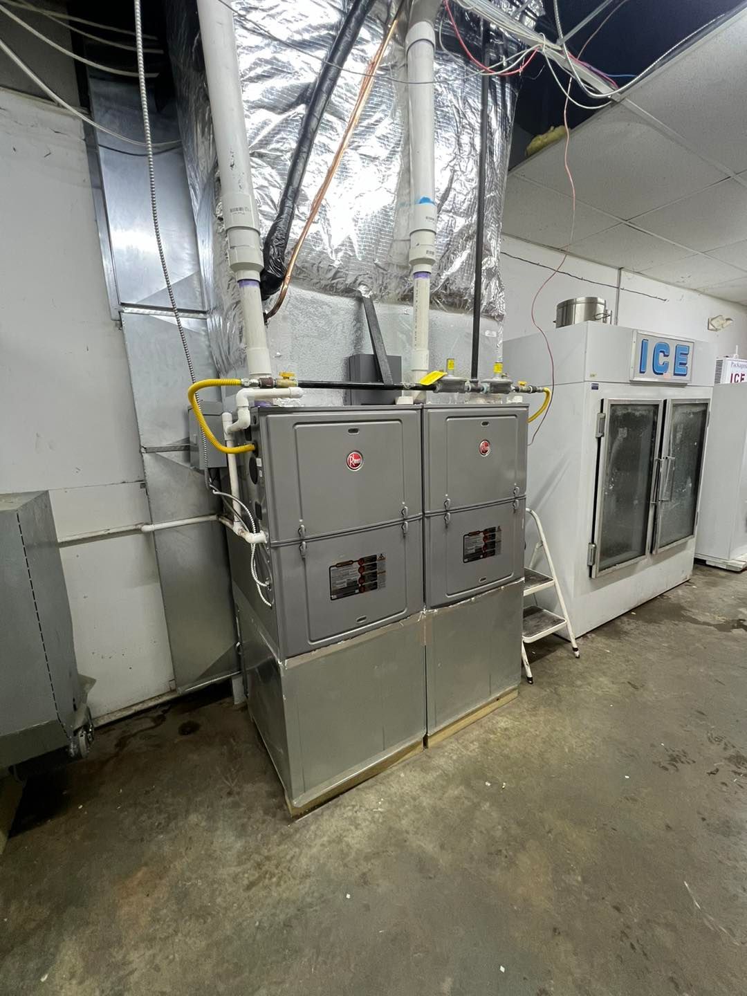 Two commercial gas furnaces installed side-by-side with silver ductwork in an industrial setting near an ice machine.