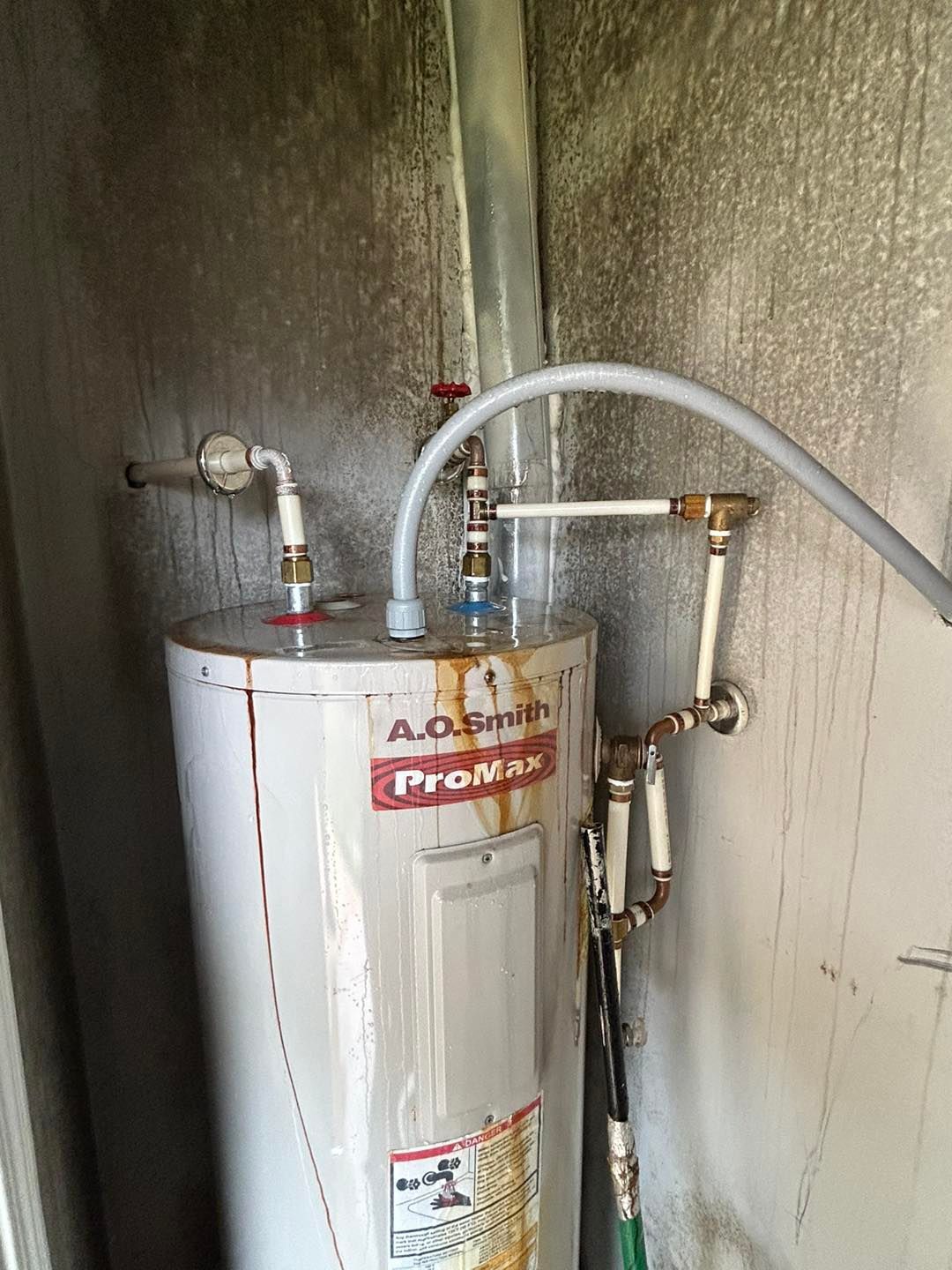 An A.O. Smith ProMax water heater with visible rust stains in a corner space with white plastic plumbing connections.