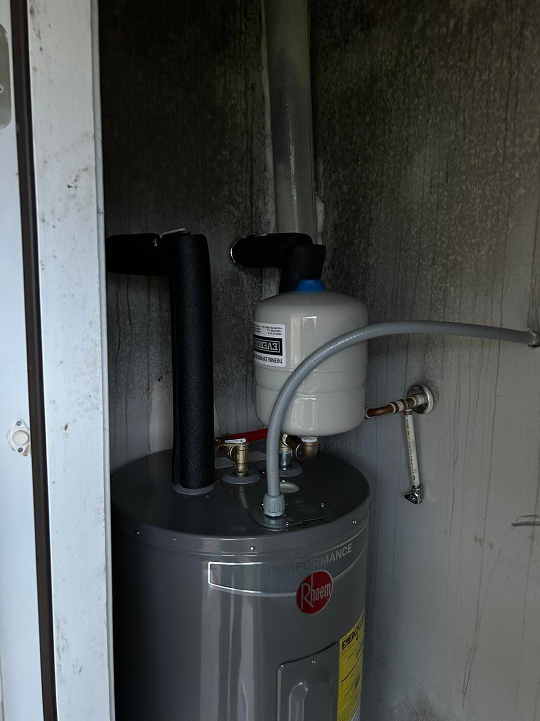 A grey Rheem water heater installed in an alcove with an expansion tank attached to the top pipes.