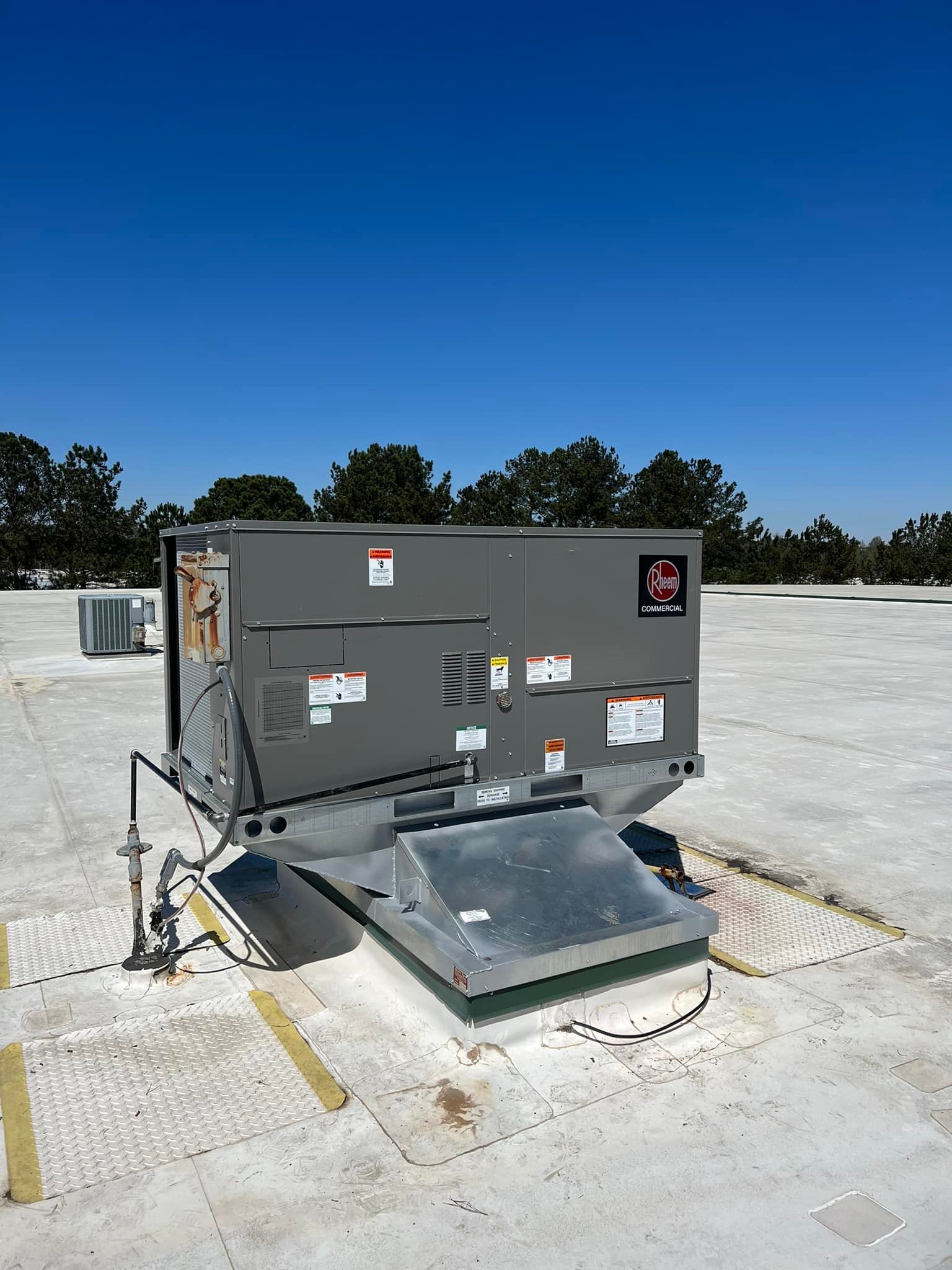 A gray rooftop HVAC unit sits on a white commercial roof against a clear blue sky.