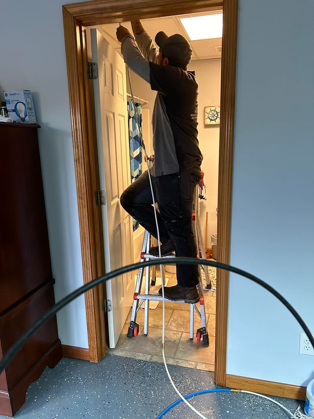 A person stands on a ladder in a doorway, working on the door frame with a long cable hanging down.
