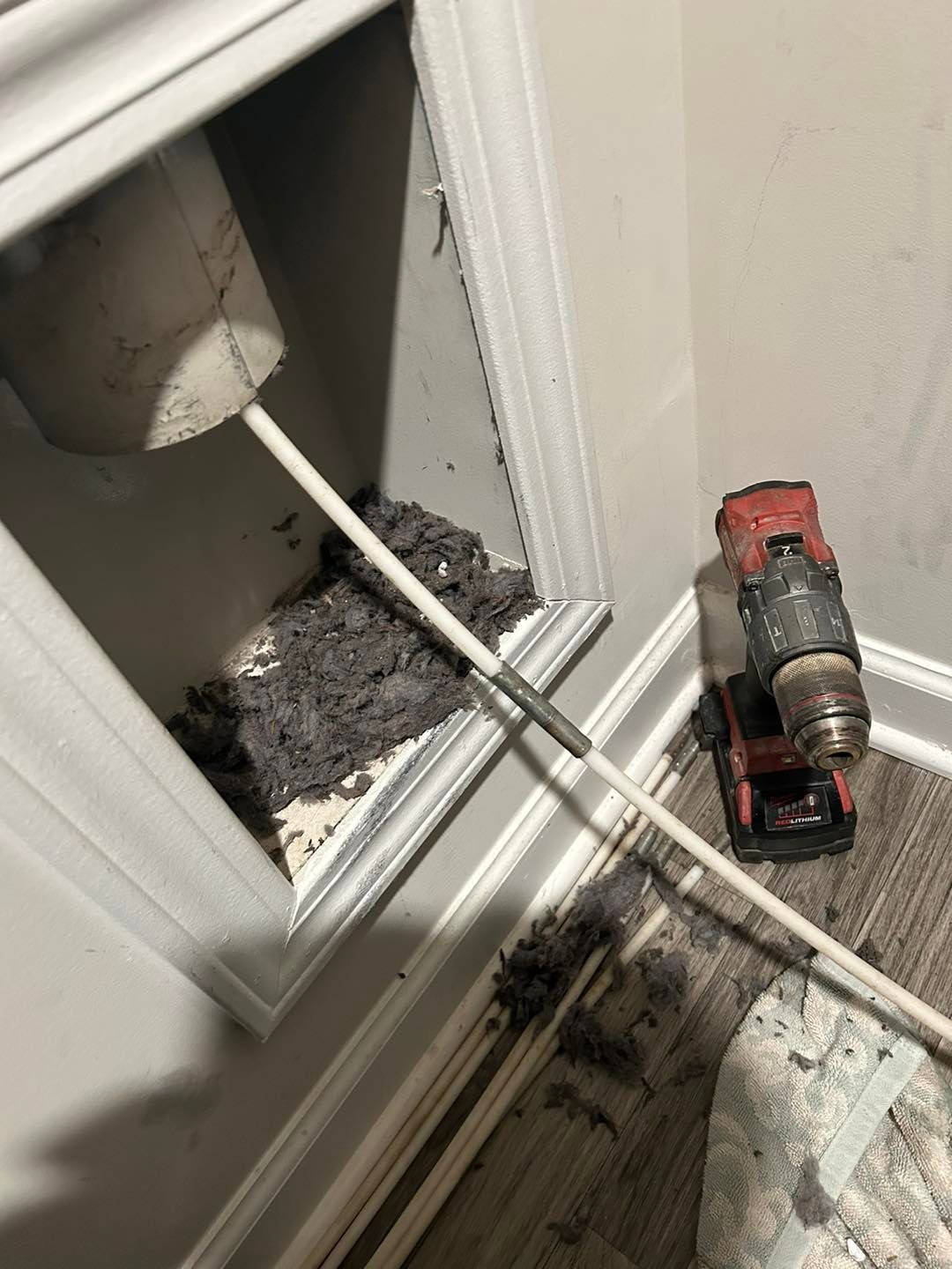 A drill with an extension brush attached, inserted into a wall cavity filled with lint to clean a dryer vent.