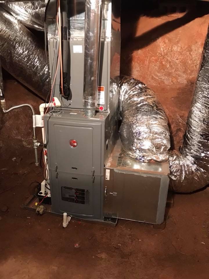 A gray HVAC furnace unit with silver ductwork installed in a dirt-floored crawl space.