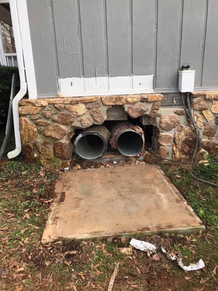 A gray-sided house exterior with a stone foundation revealing a large opening with two exposed metal pipes.