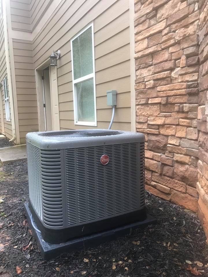 A grey Rheem air conditioning unit sits on a black pad next to a house with beige siding and stone accents.
