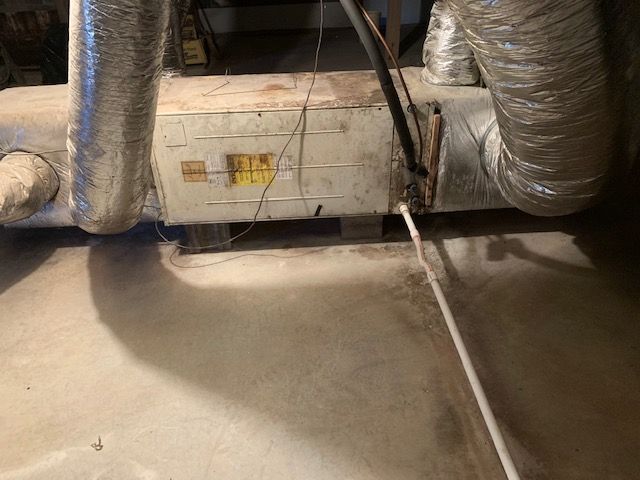 An HVAC air handler unit with silver flexible ductwork and a white PVC drain line situated on a concrete floor.