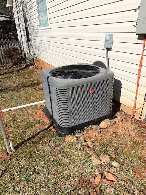 A grey residential outdoor HVAC condenser unit sits on a dirt and rock patch against a house with light beige siding.