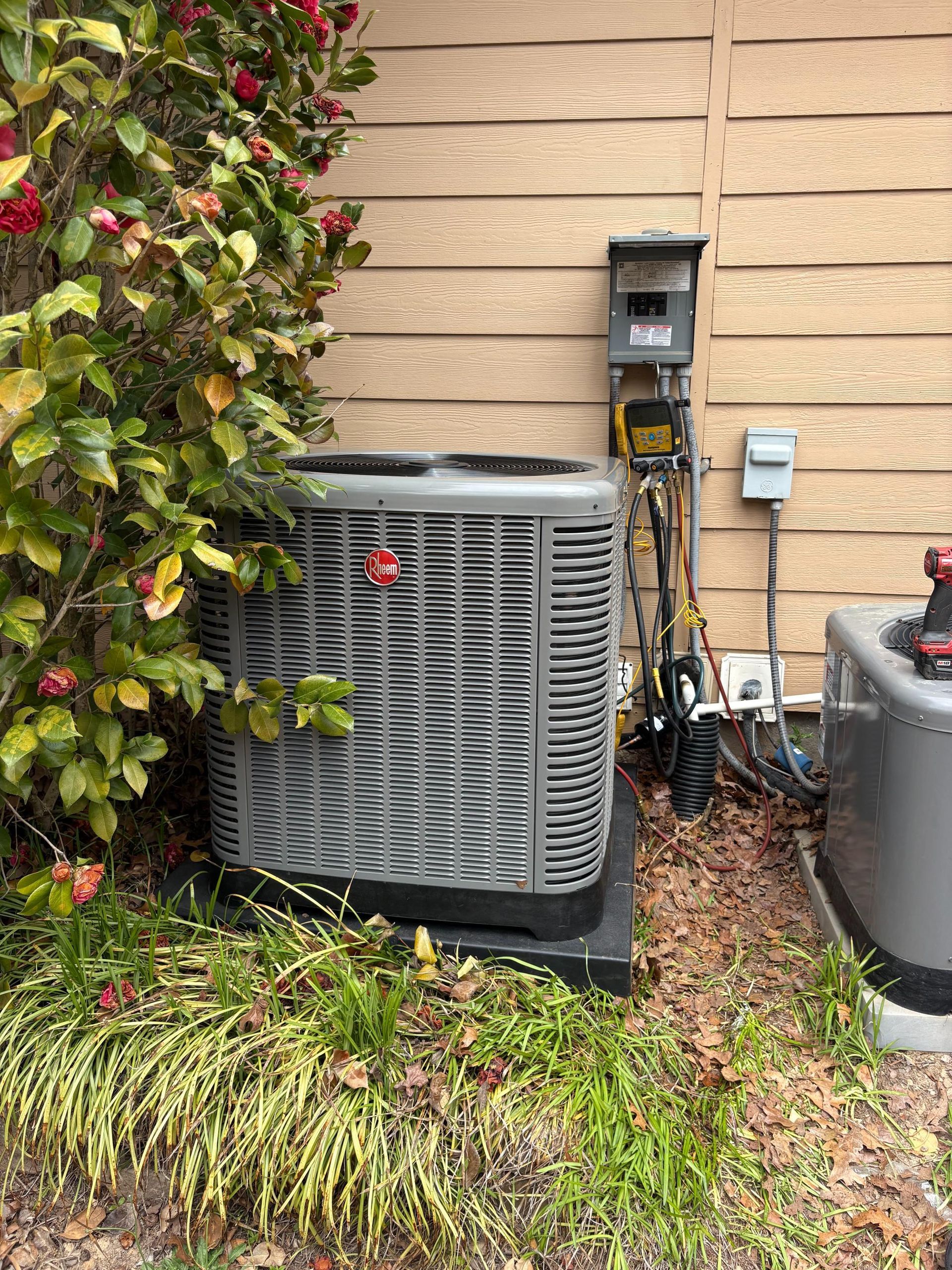 Outdoor air conditioner unit beside a house wall with plants and utility boxes nearby
