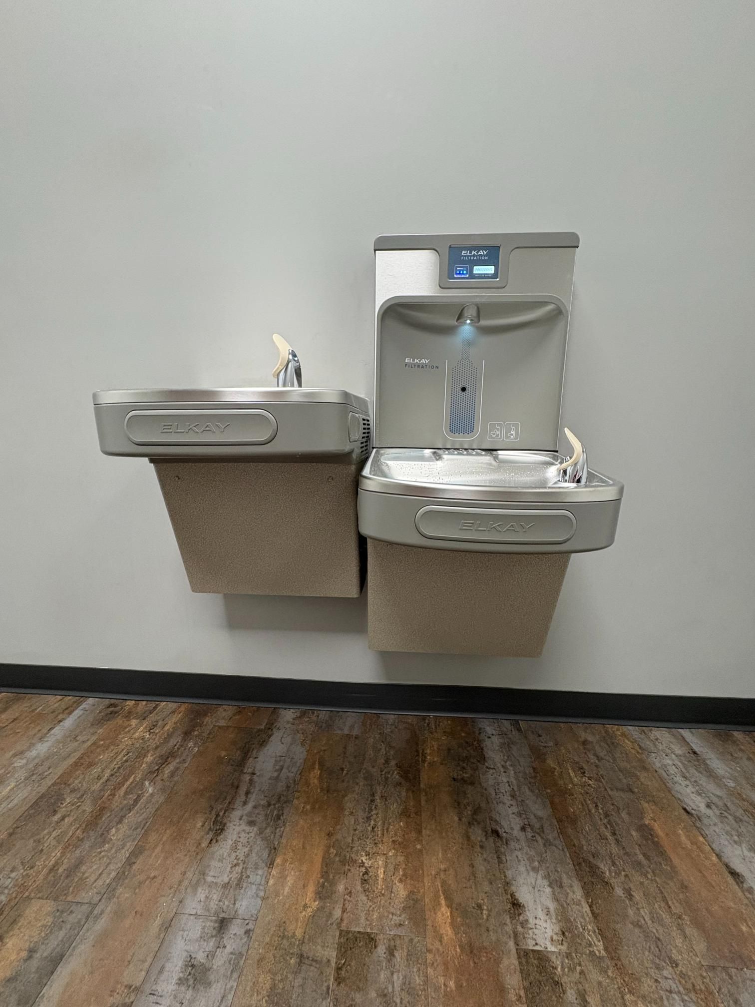 Two stainless steel drinking fountains mounted on a wall above a wood-look floor.