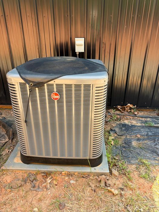 A smiling person in a uniform holds a young child while standing next to a grey Rheem HVAC unit against a stone wall.