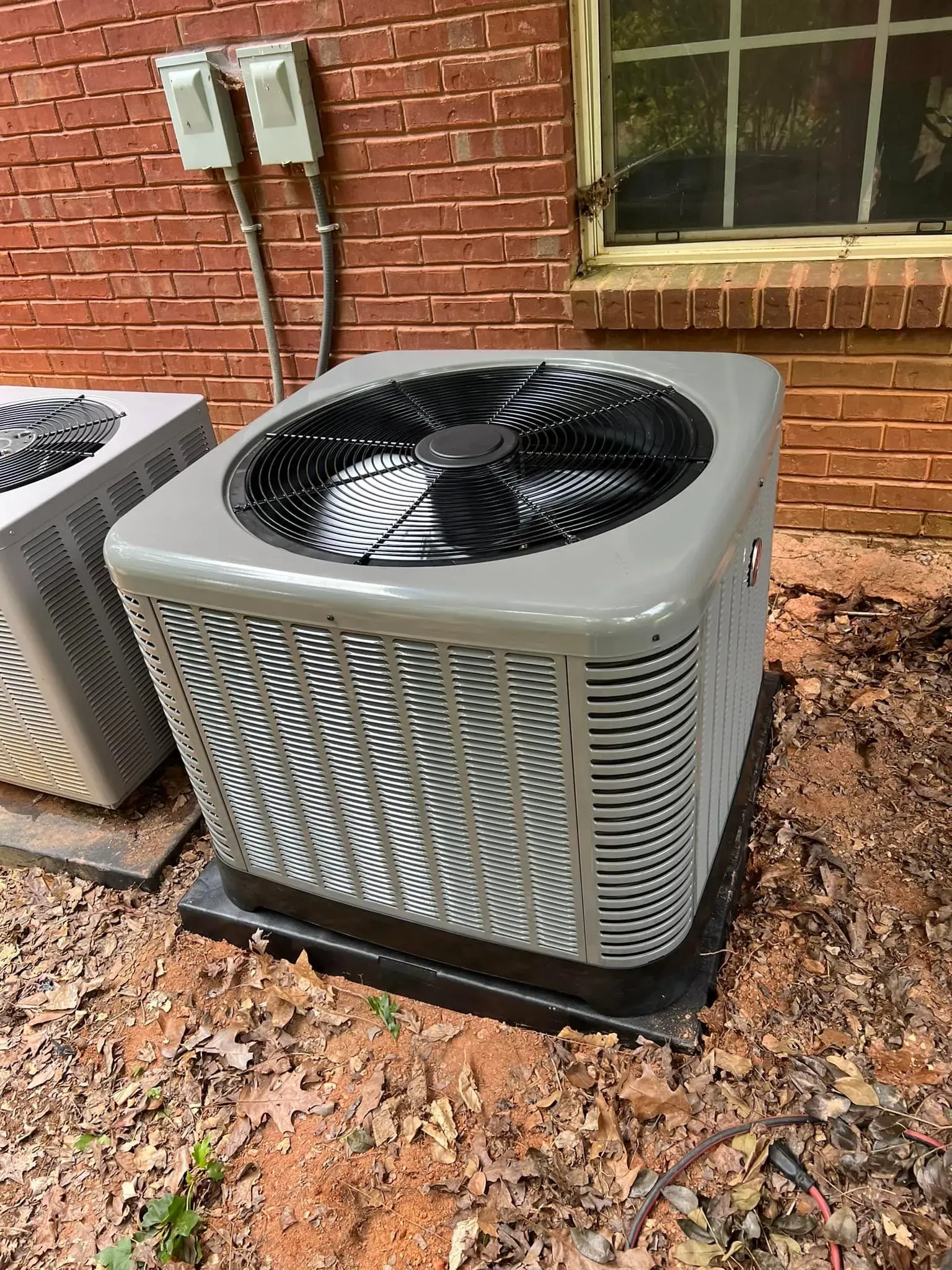A toddler in a baseball cap and tan pants pretends to work on a large grey Rheem HVAC unit against a stone wall.