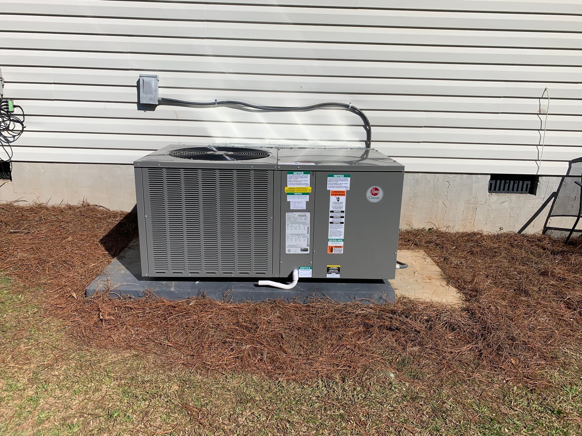A gray outdoor air conditioning unit sits on a concrete pad against the light-colored vinyl siding of a house.