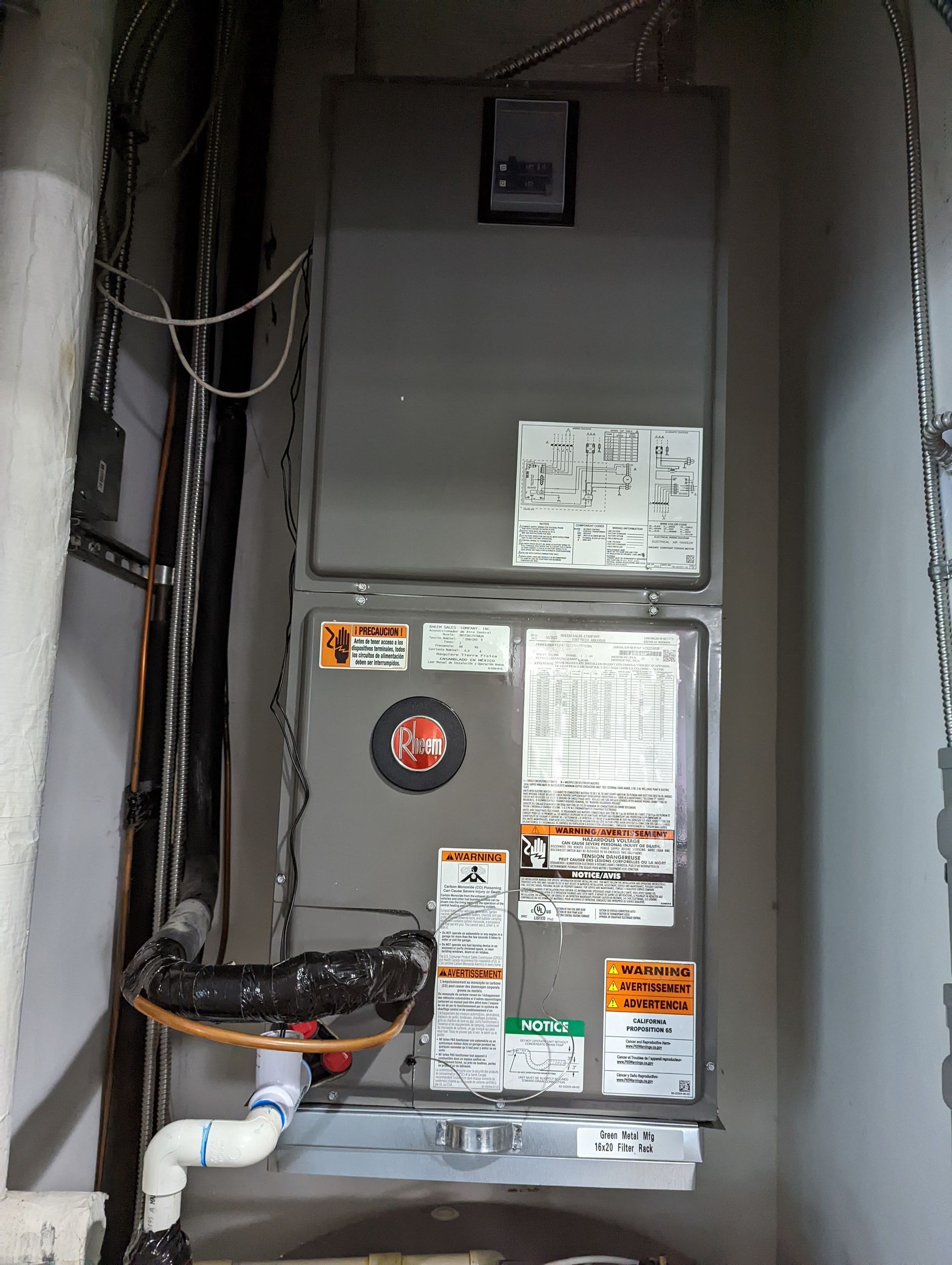 A vertical grey HVAC unit mounted on a wall with white drain pipes and various safety labels.