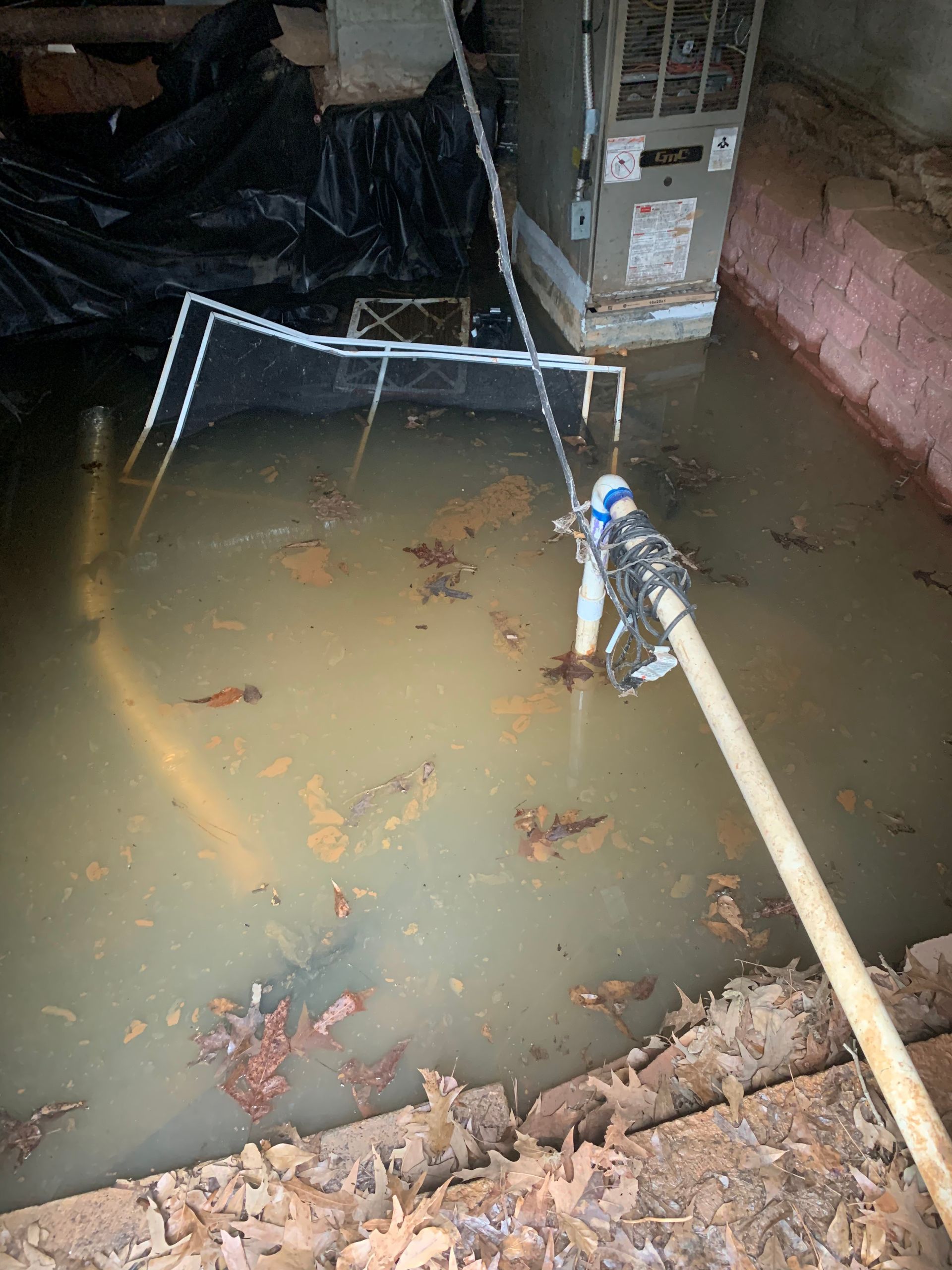 A flooded crawl space containing standing water, debris, an HVAC unit, and a drainage pump with a hose.