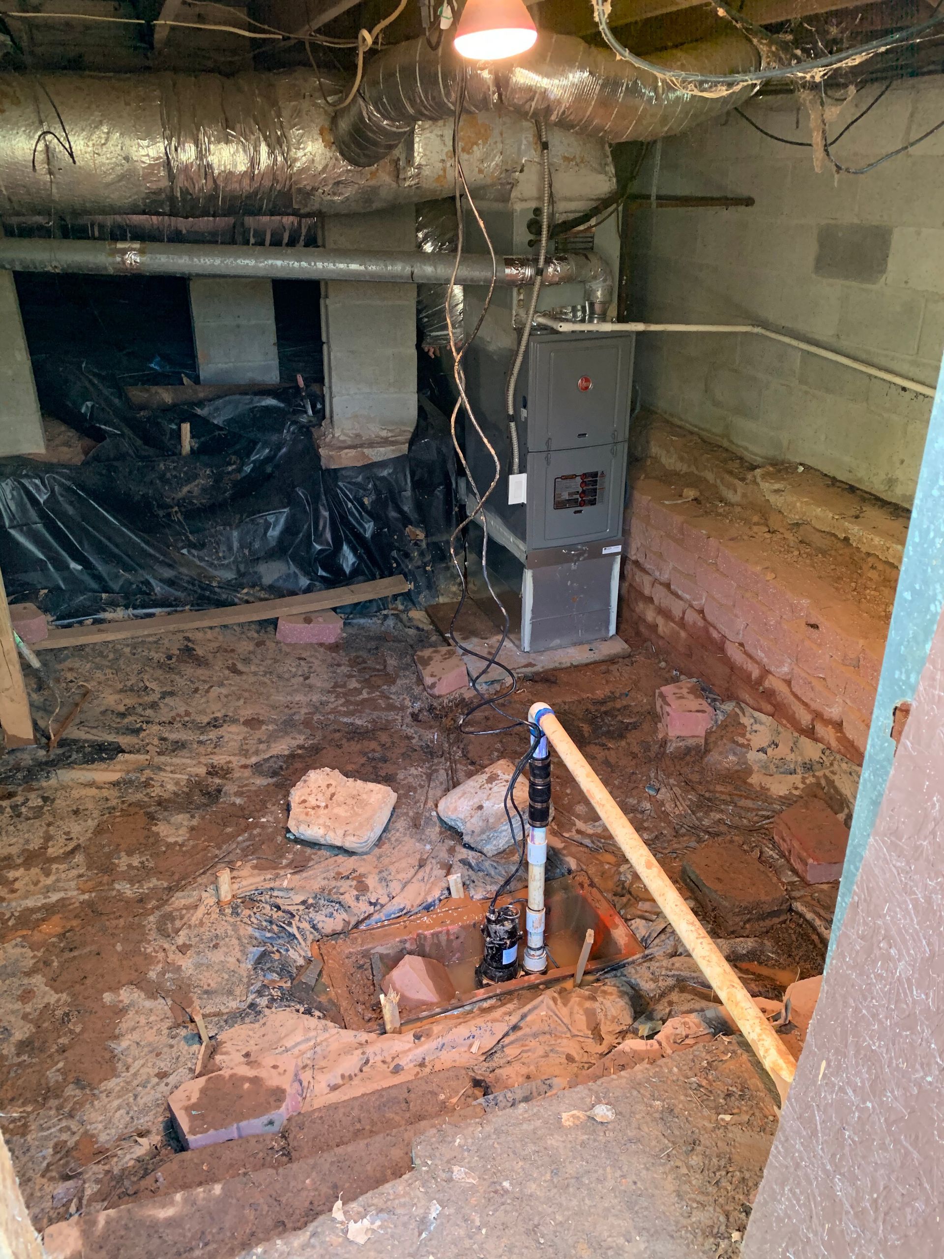 A dimly lit crawl space with exposed soil, a furnace, ductwork, a plastic vapor barrier, and a sump pump system.