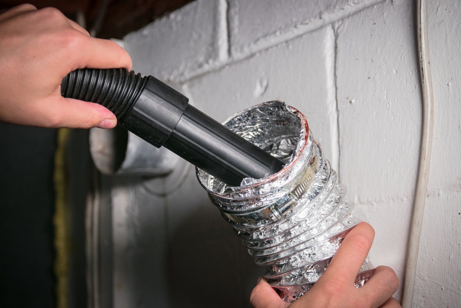Hands use a vacuum hose attachment to clean out a flexible aluminum dryer vent pipe mounted to a white cinder block wall.