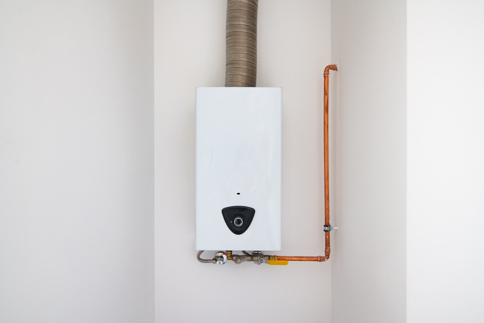 A white tankless water heater mounted on a white wall with copper piping and an exhaust vent extending upward.