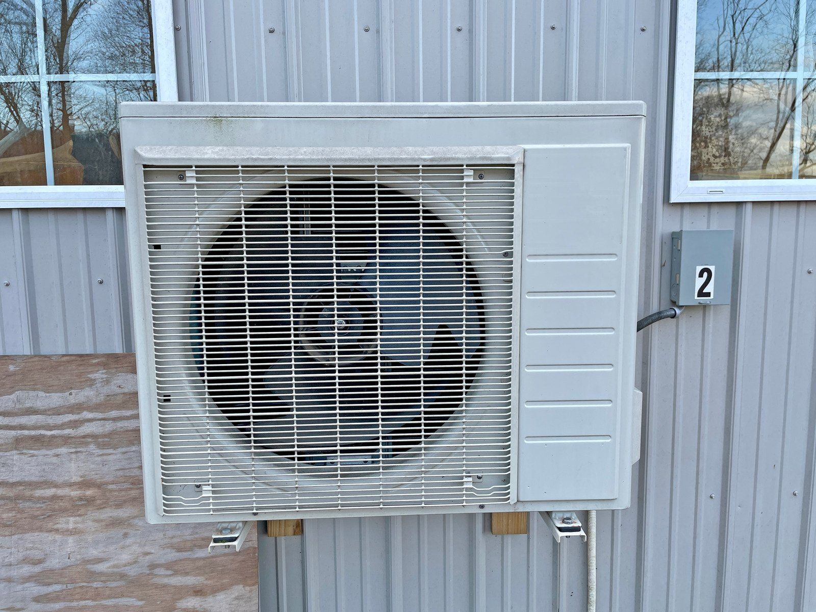 Light gray outdoor air conditioning condenser unit mounted on a corrugated metal wall with a labeled electrical box.