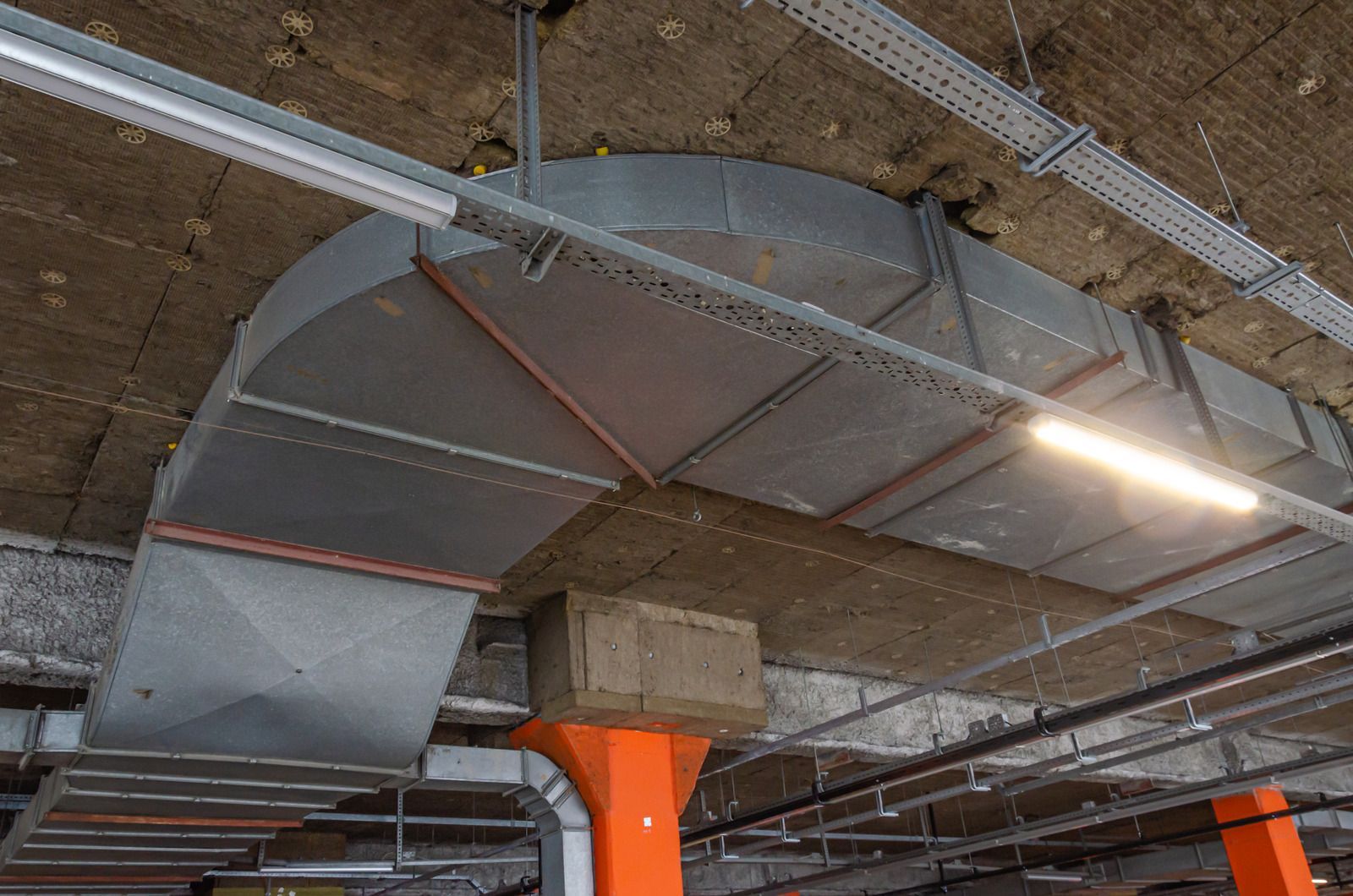 Large metal ventilation ducts run along a dark concrete ceiling near bright orange painted support columns.