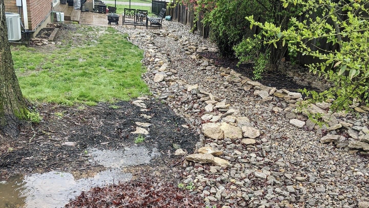 Rocky garden path with muddy puddles, bordered by grass and bushes beside a brick wall