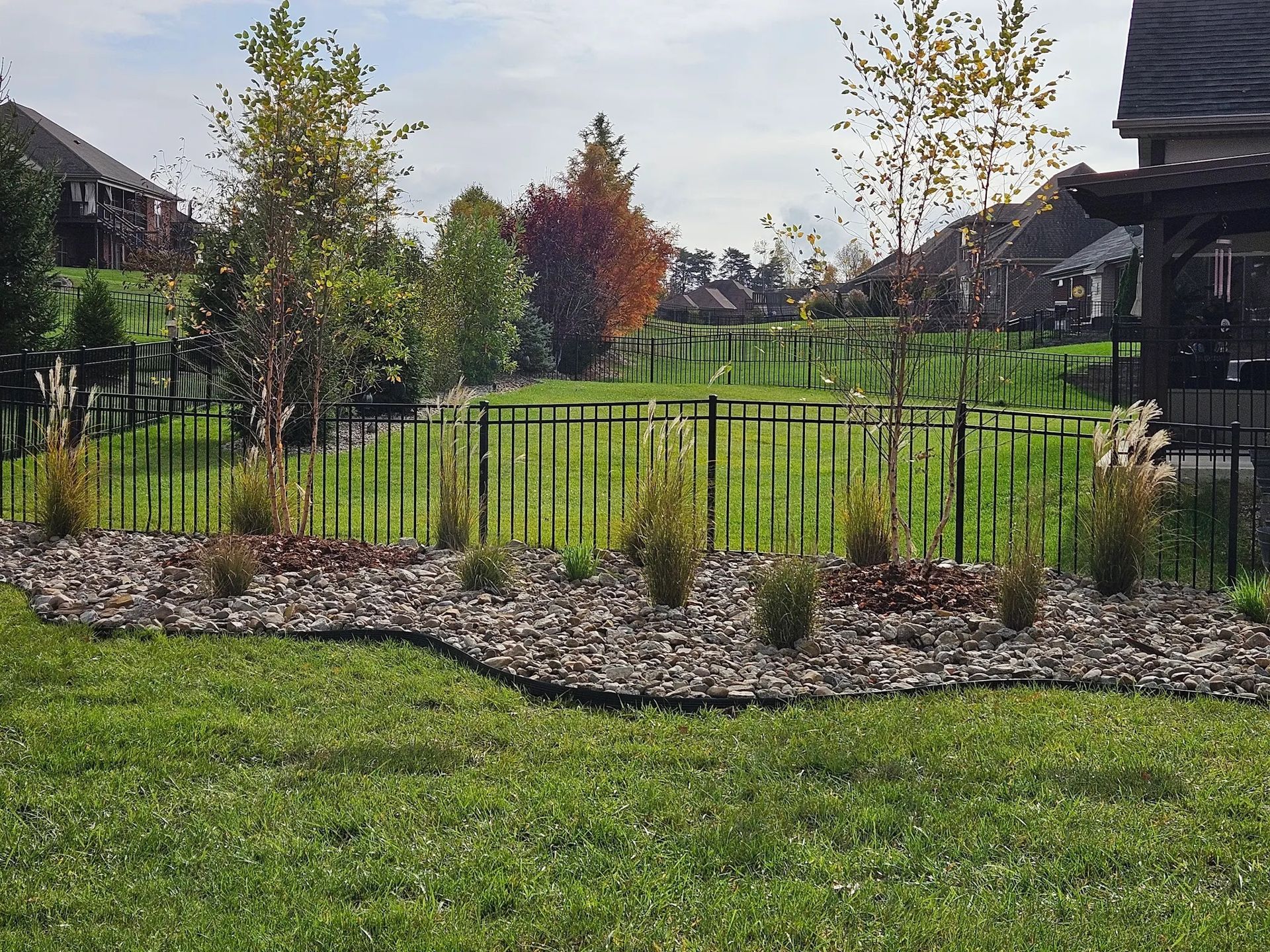 Black metal fence encloses a landscaped yard with mulch, plants, and trees. Green grass in the foreground.