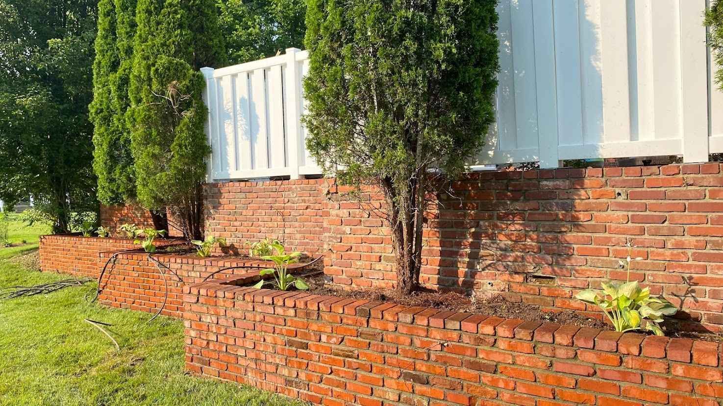 Brick retaining wall with green shrubs beneath a white fence in bright sunlight