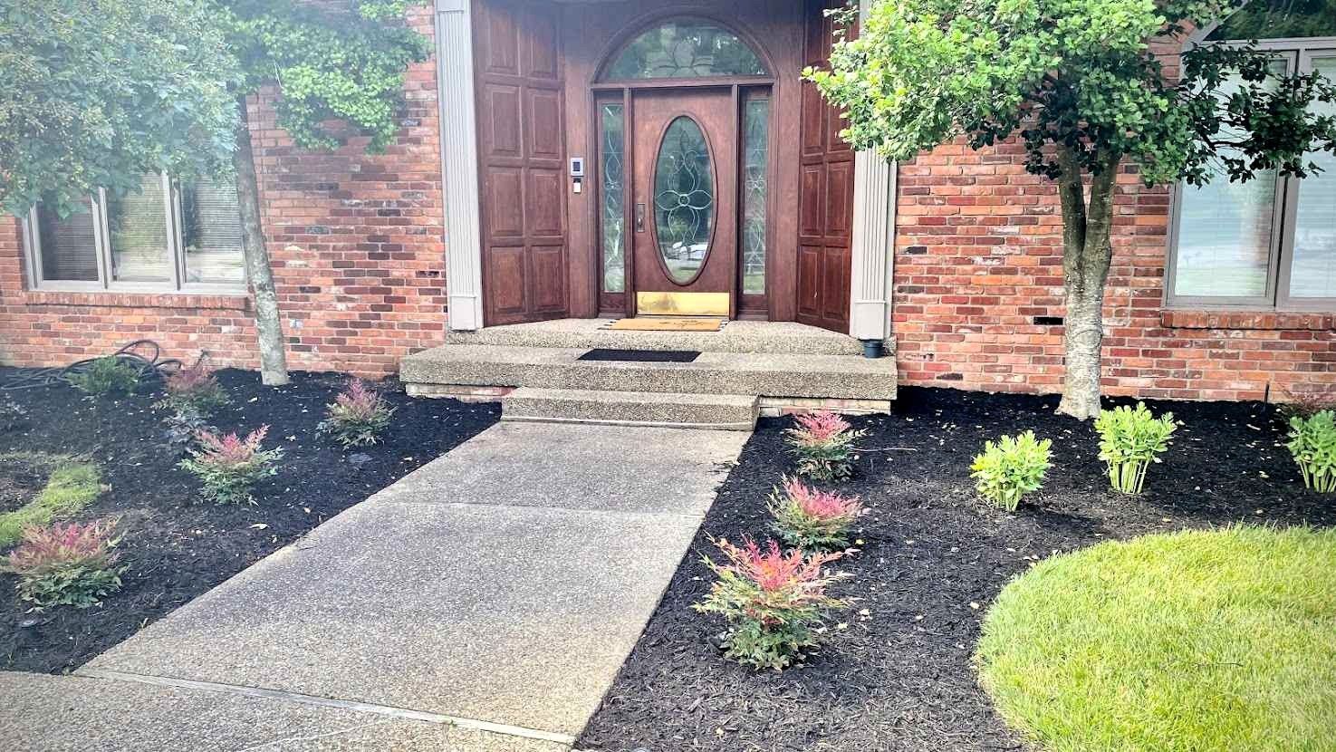 A brick house with a concrete walkway leading to a wooden door, flanked by trees and flower beds.
