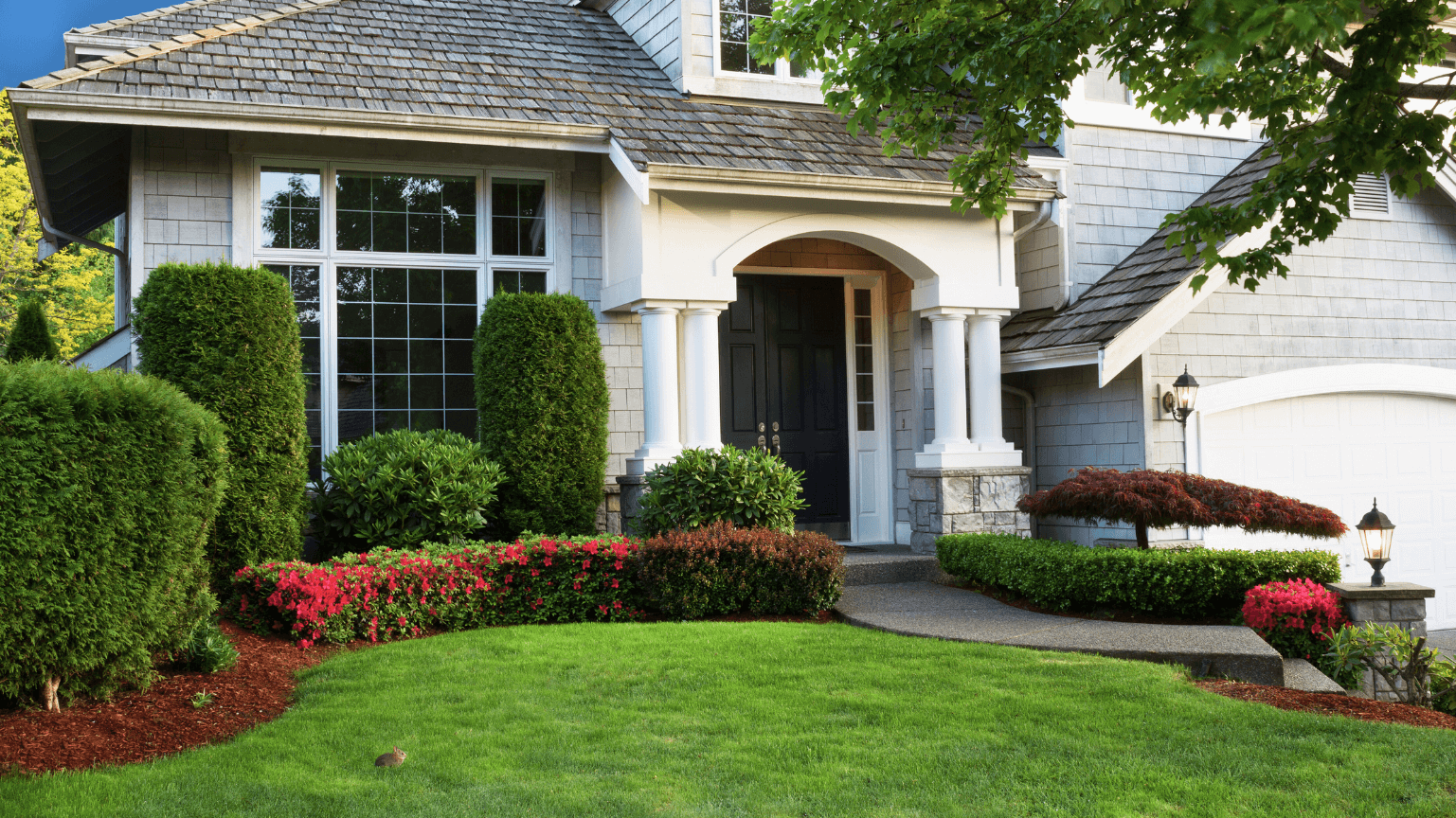 Gray house with well-manicured green lawn and landscaping, including shrubs, bushes, and red flowers.