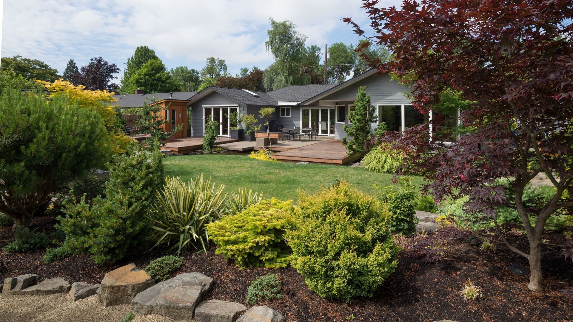A gray house with a wraparound deck surrounded by a landscaped garden with green and yellow bushes, trees, and a green lawn.