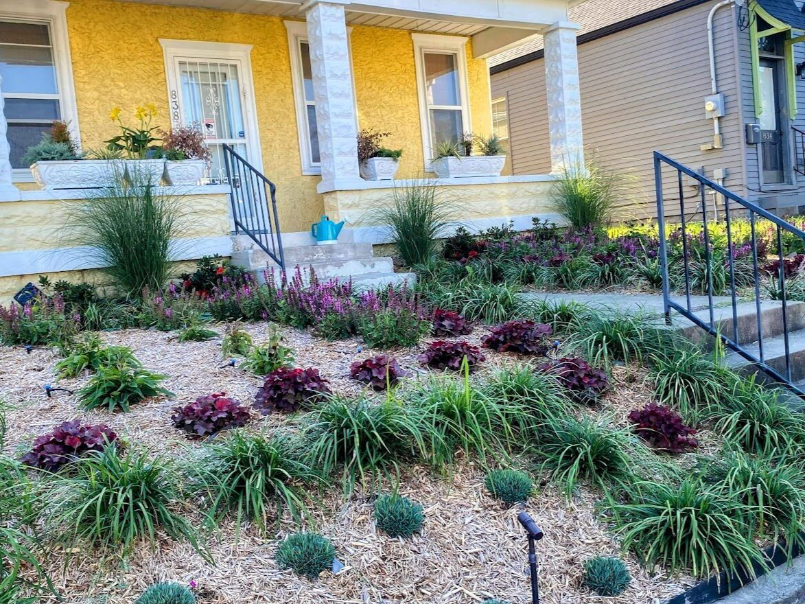 Front porch with stone steps, black railings, and landscaped flower beds in front of a brick house