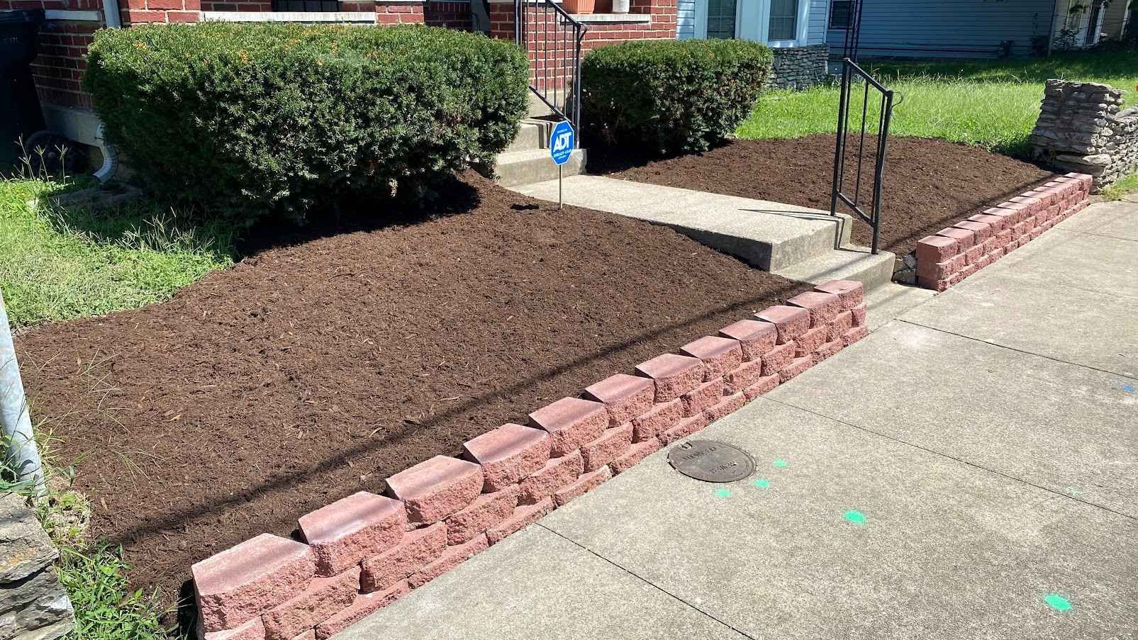 A brick-lined flowerbed filled with mulch leads to a home's steps and entrance, with green bushes on either side.