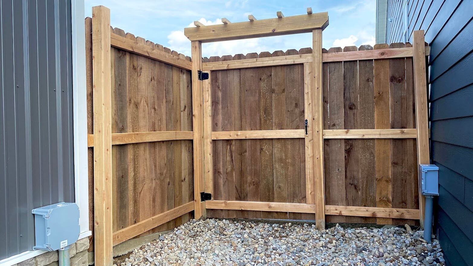 Wooden backyard fence with a gate, gravel ground, and neighboring buildings in the background