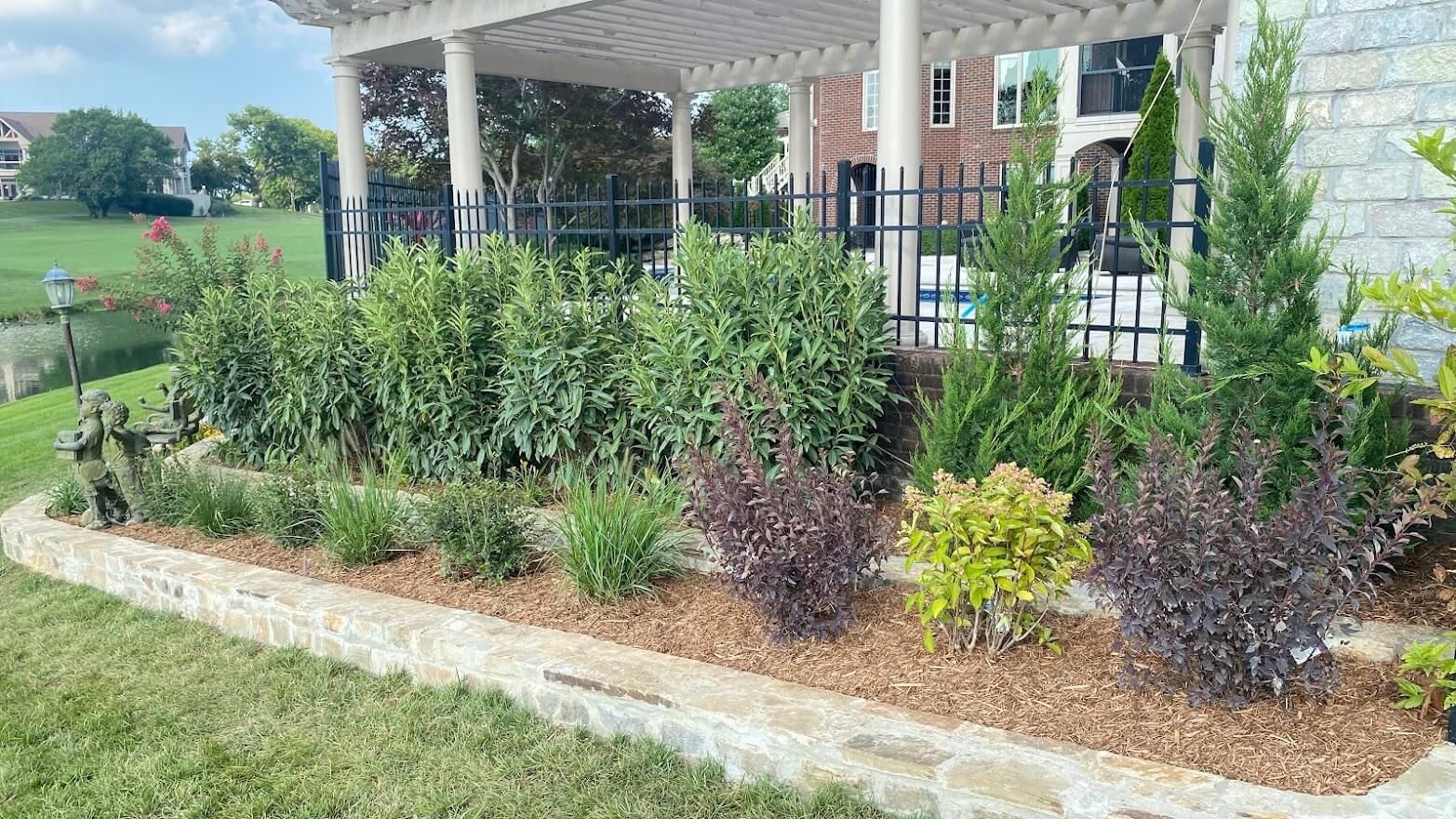 Landscaped garden bed with various shrubs and flowers, a pergola in the background, and a lake on a sunny day.
