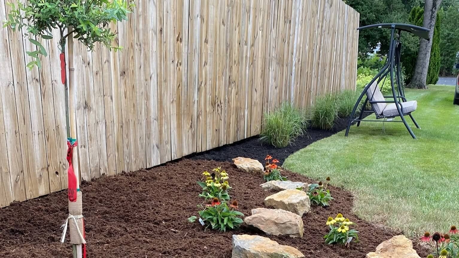Mulched garden bed with rocks and young plants beside a wooden fence and lawn swing.