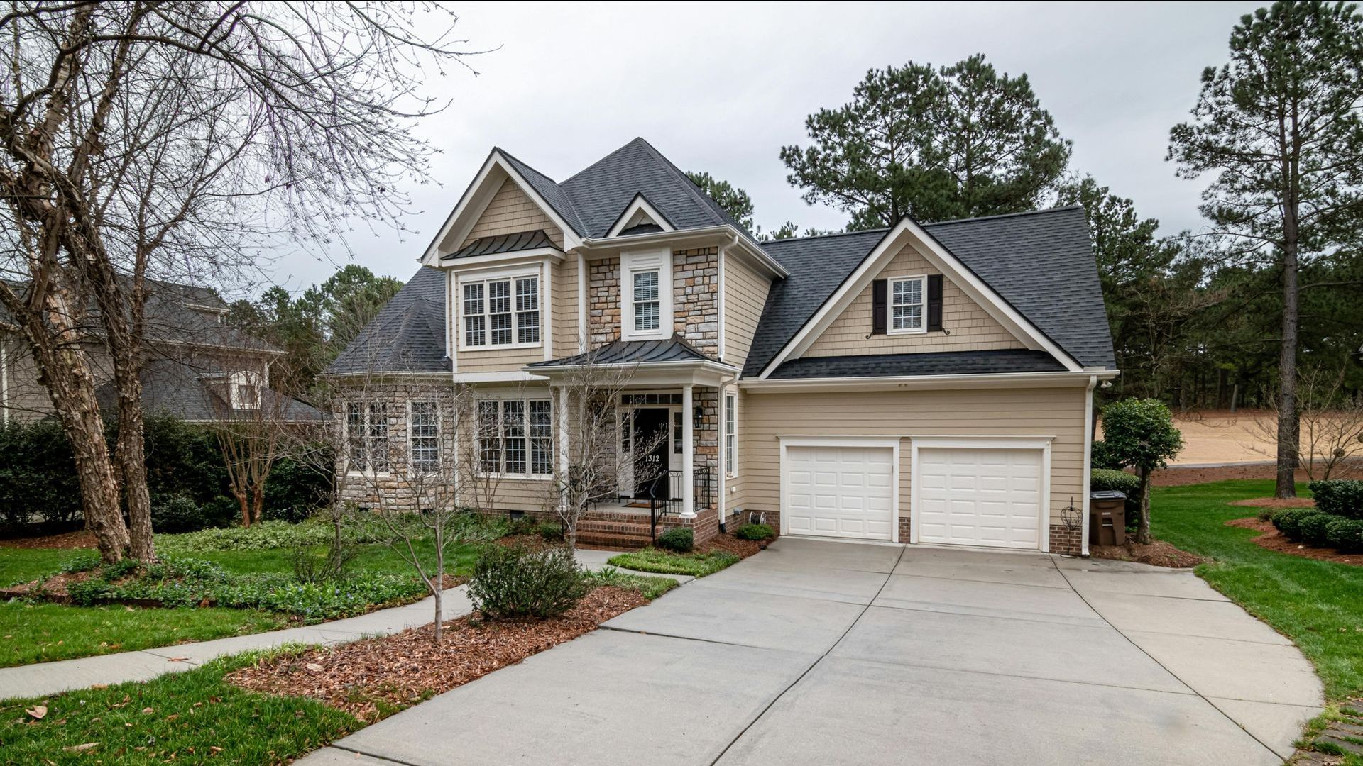 Two-story beige house with gray roof, stone accents, and a two-car garage. Landscaped front yard with driveway.