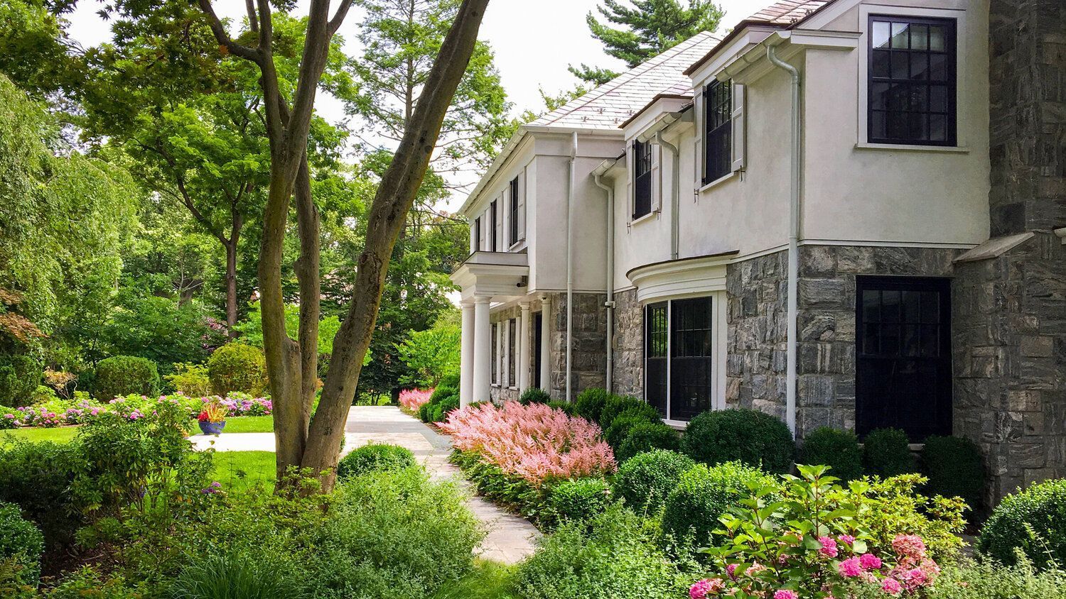 Exterior of a stately, white two-story house with stone accents, surrounded by lush green gardens and pink flowers.