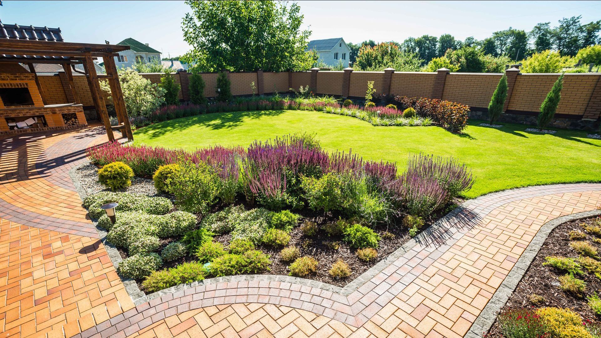A lush backyard with a brick path, lawn, flower beds, wooden fence, and pergola.