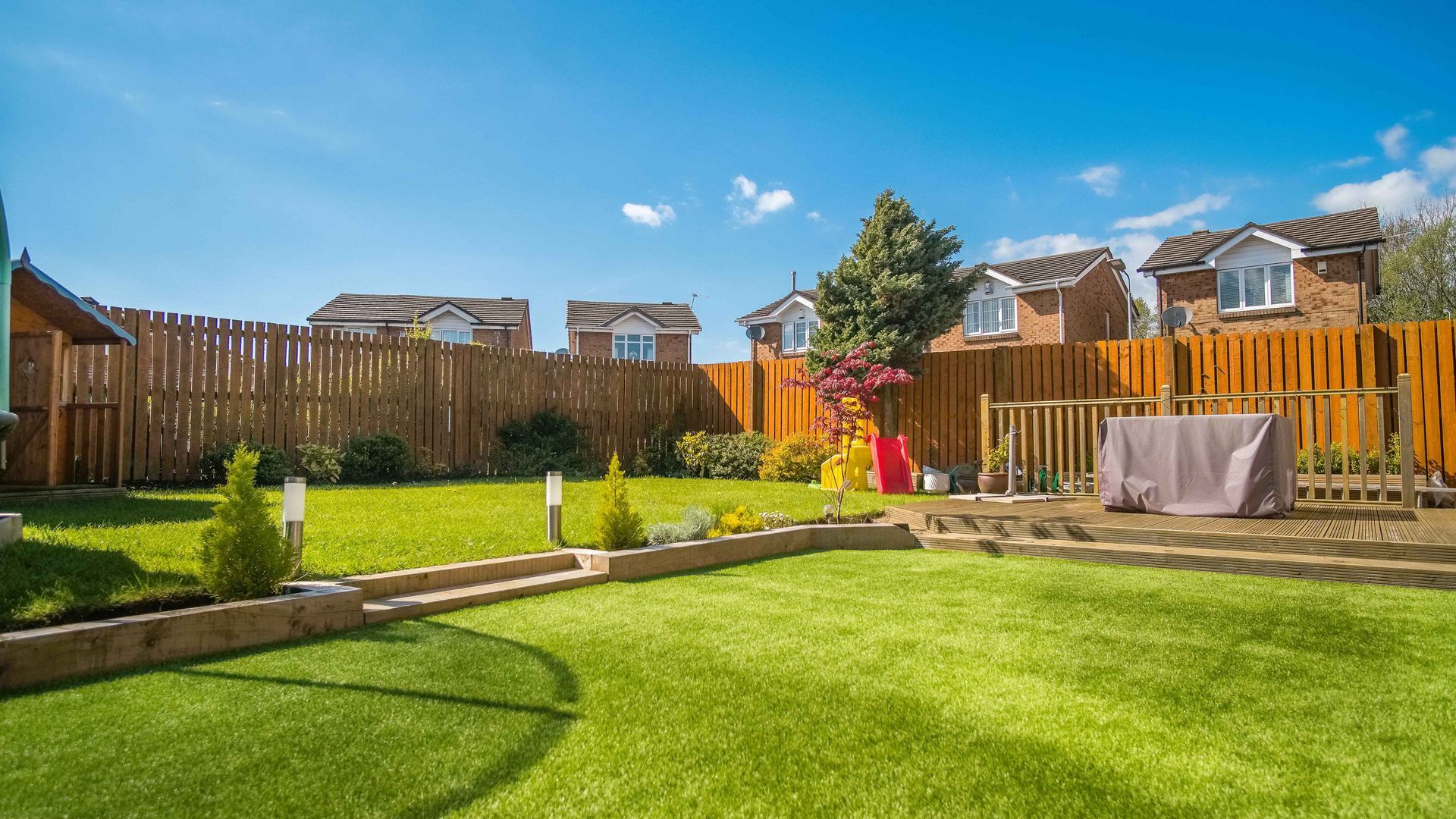 Green backyard with manicured lawn, wooden fence, and houses under a bright blue sky.