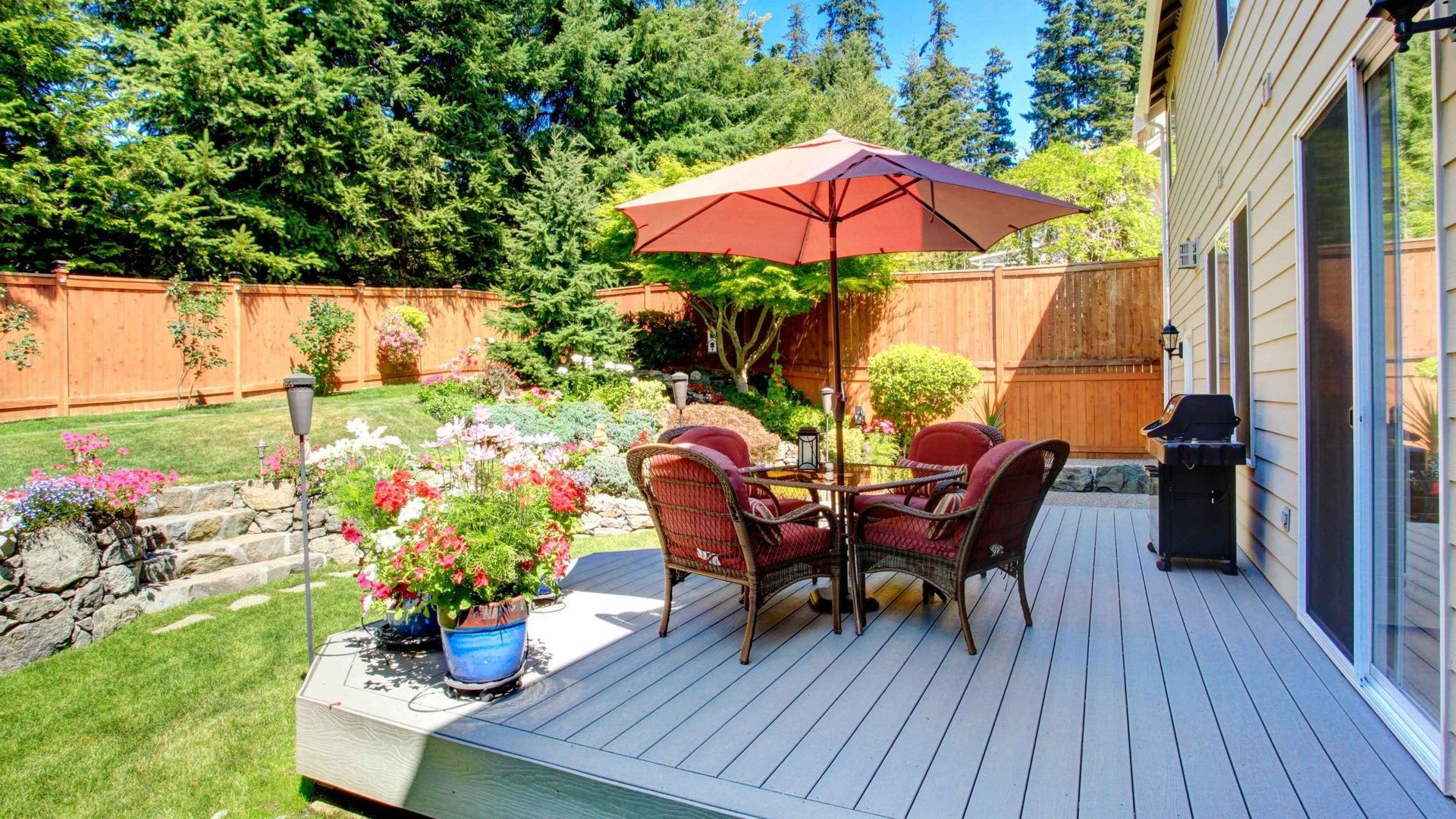 Backyard patio with a table, chairs, and umbrella on a wooden deck, with greenery, flowers, and a fence.