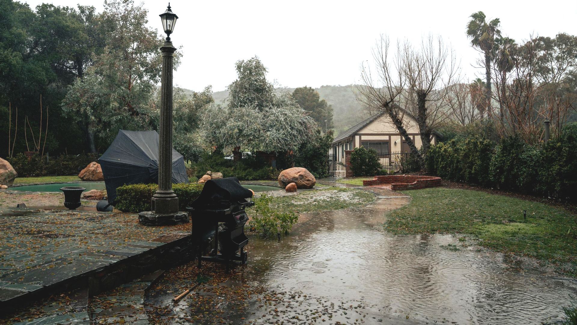 A flooded yard with a home in the background. An open black umbrella and a lamppost are in the yard.