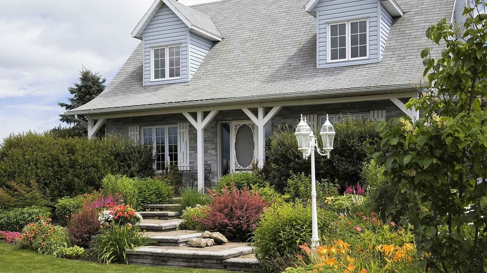 A light blue house with white trim, dormers, and a porch, surrounded by a colorful garden and stone steps.