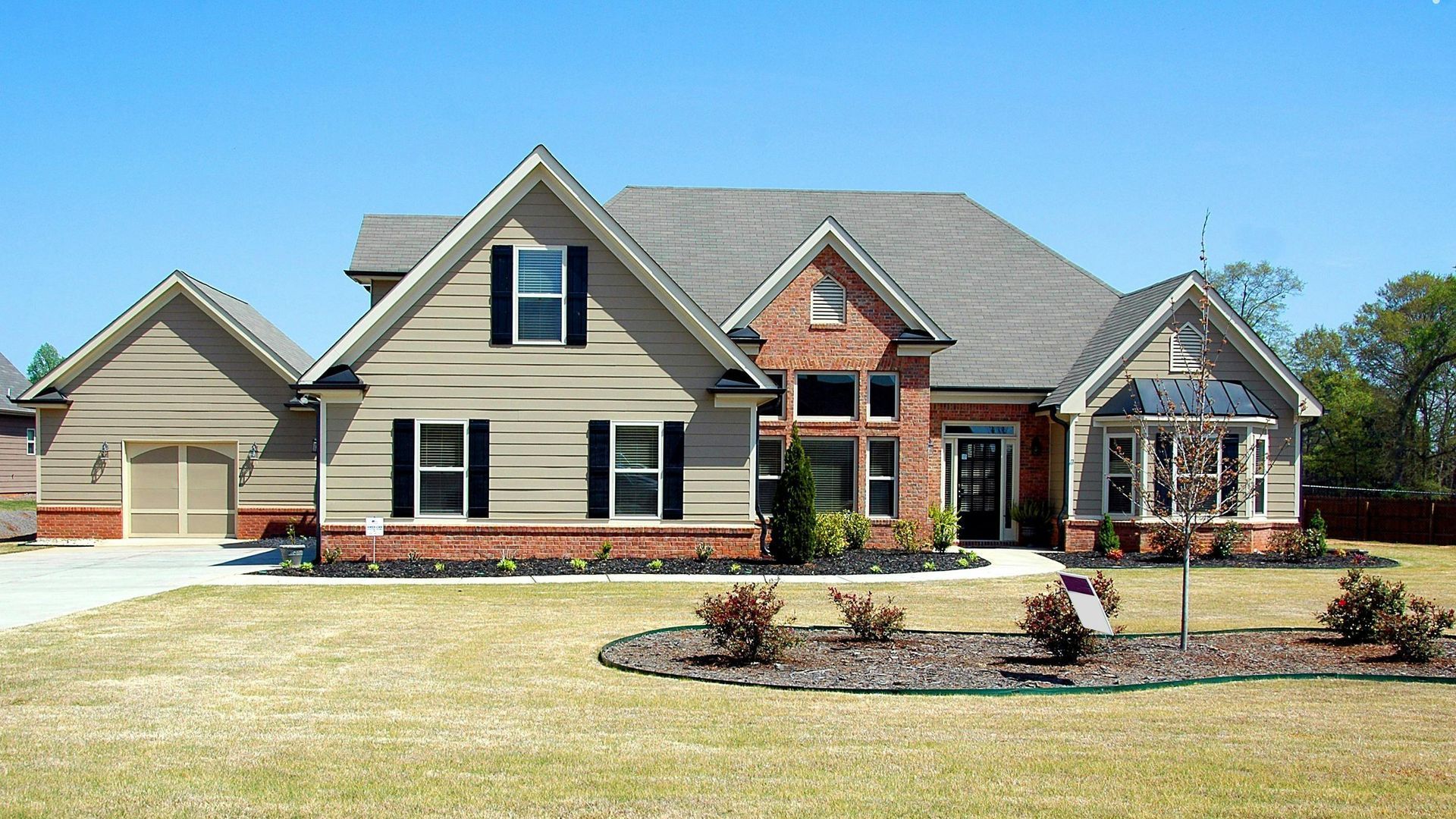 Two-story house with beige siding, brick accents, and a manicured lawn under a clear blue sky.