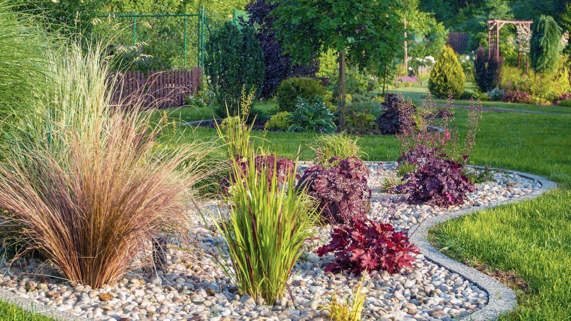 Garden bed with various colorful plants and grasses surrounded by gravel and bordered by a curved concrete edge.