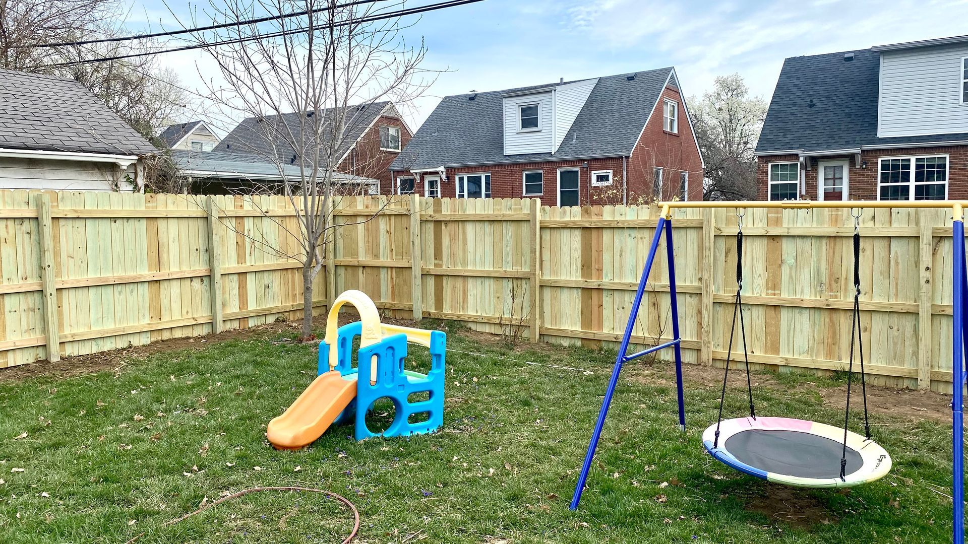 Backyard play area with a plastic slide, swing set, and trampoline inside a fenced yard.