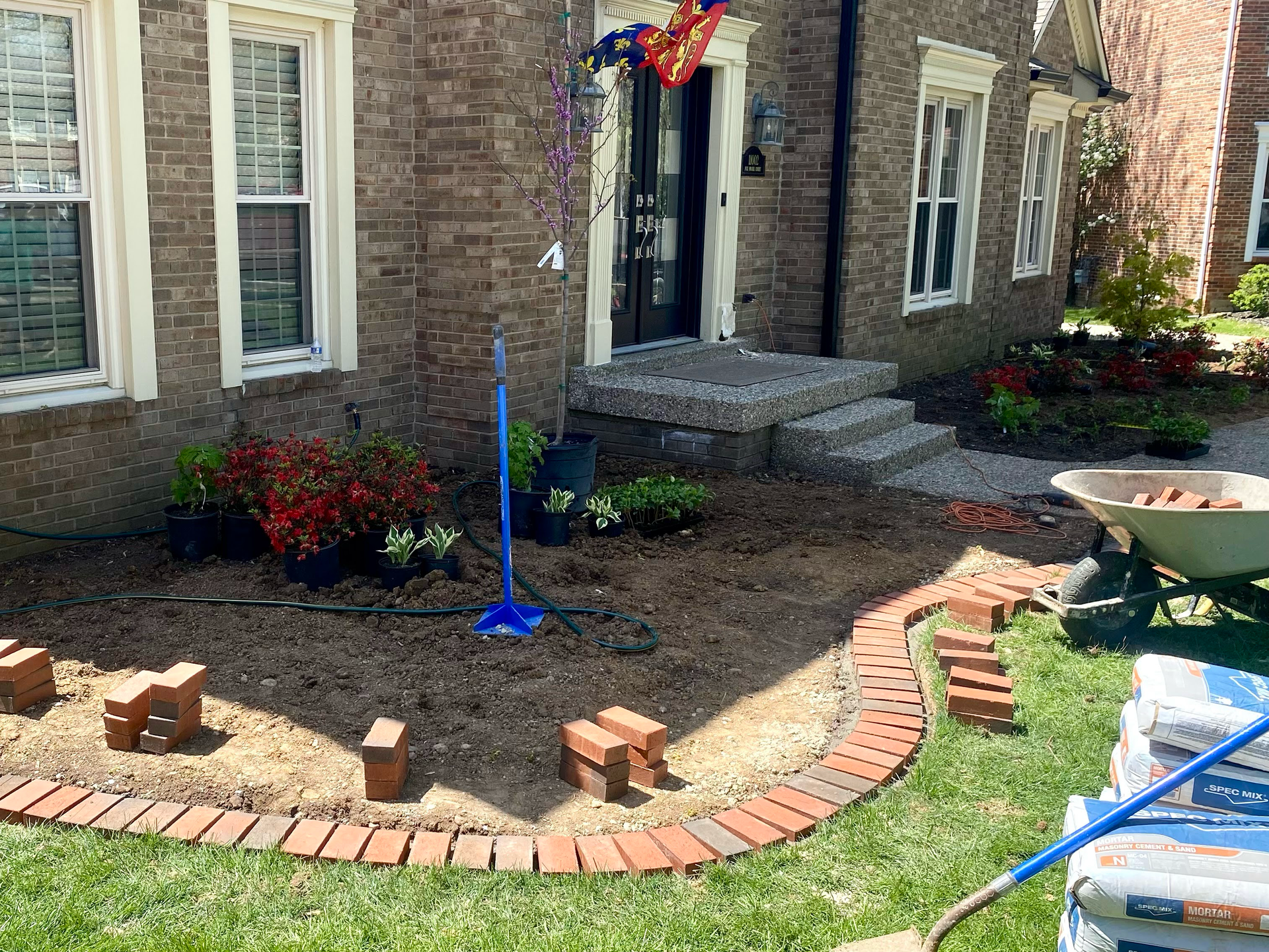 Front yard landscaping with curved brick edging, small plants, and a blue flagpole near a brick house entrance