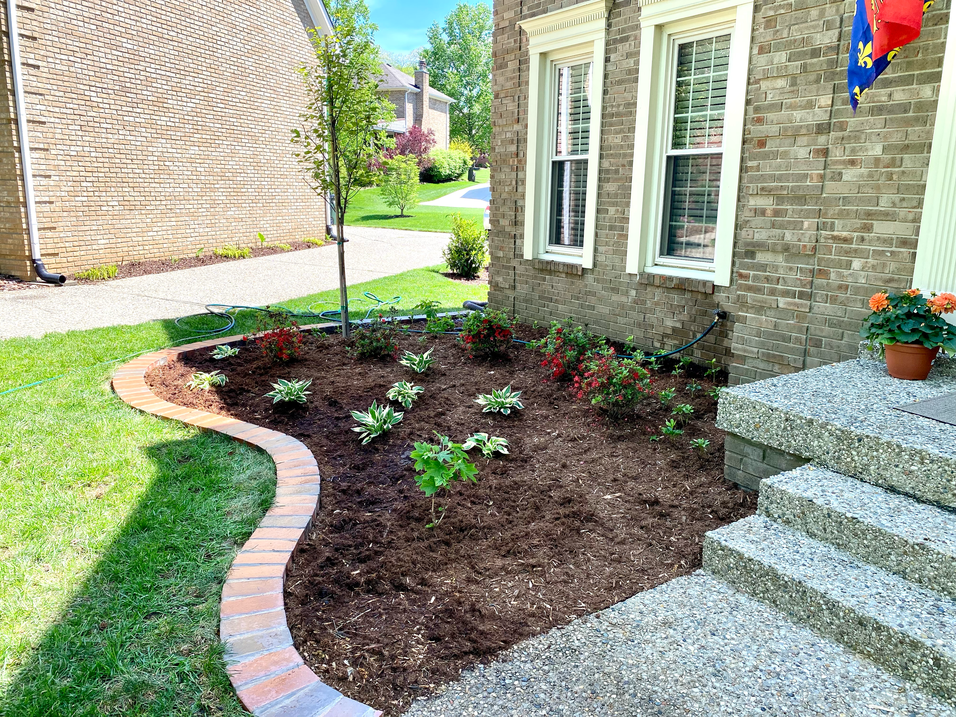 Front yard flower bed with dark mulch, small plants, brick edging, and house steps beside a brick home