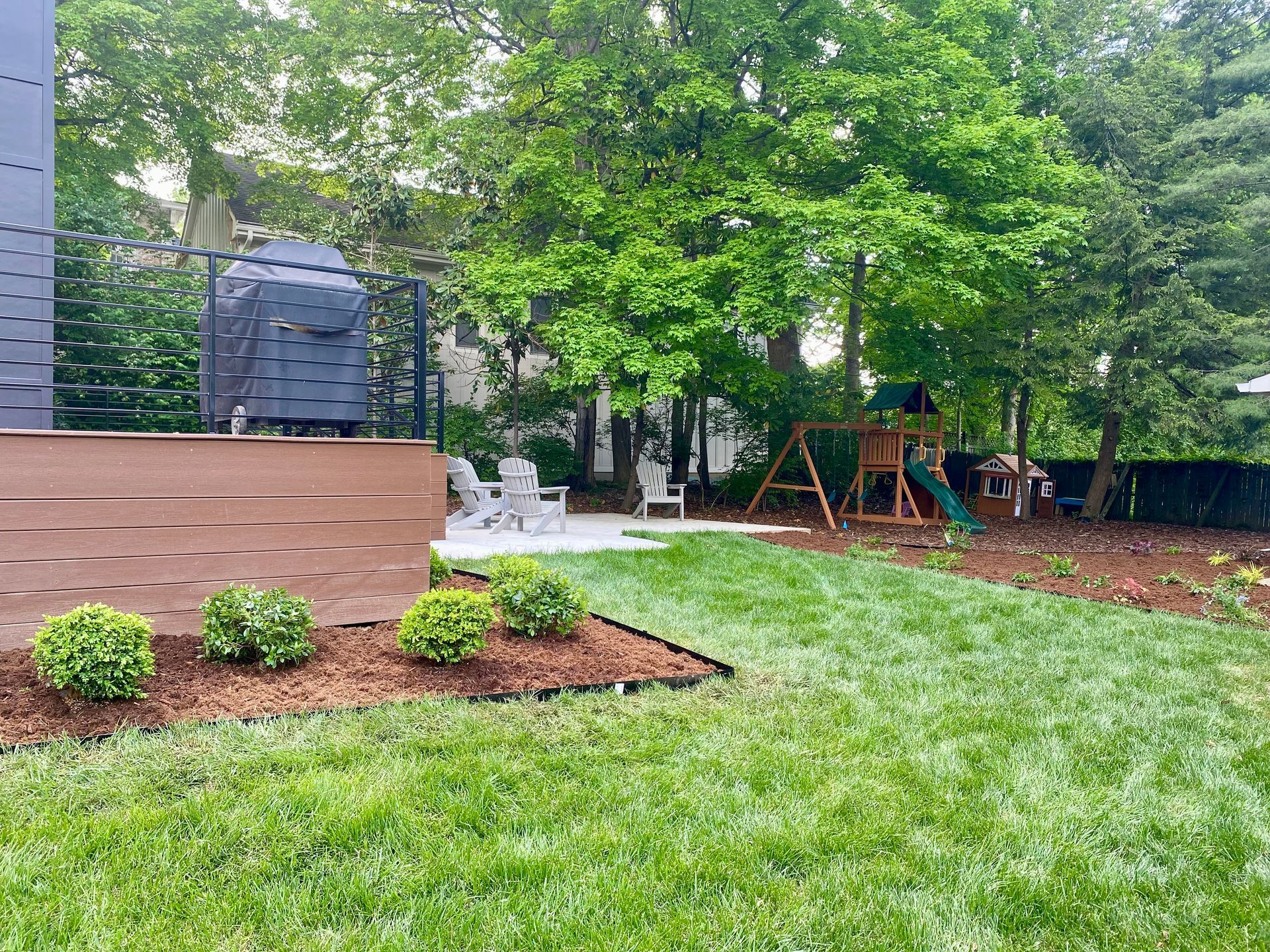 Backyard patio with wooden deck, green lawn, trees, and a swing set under overcast skies