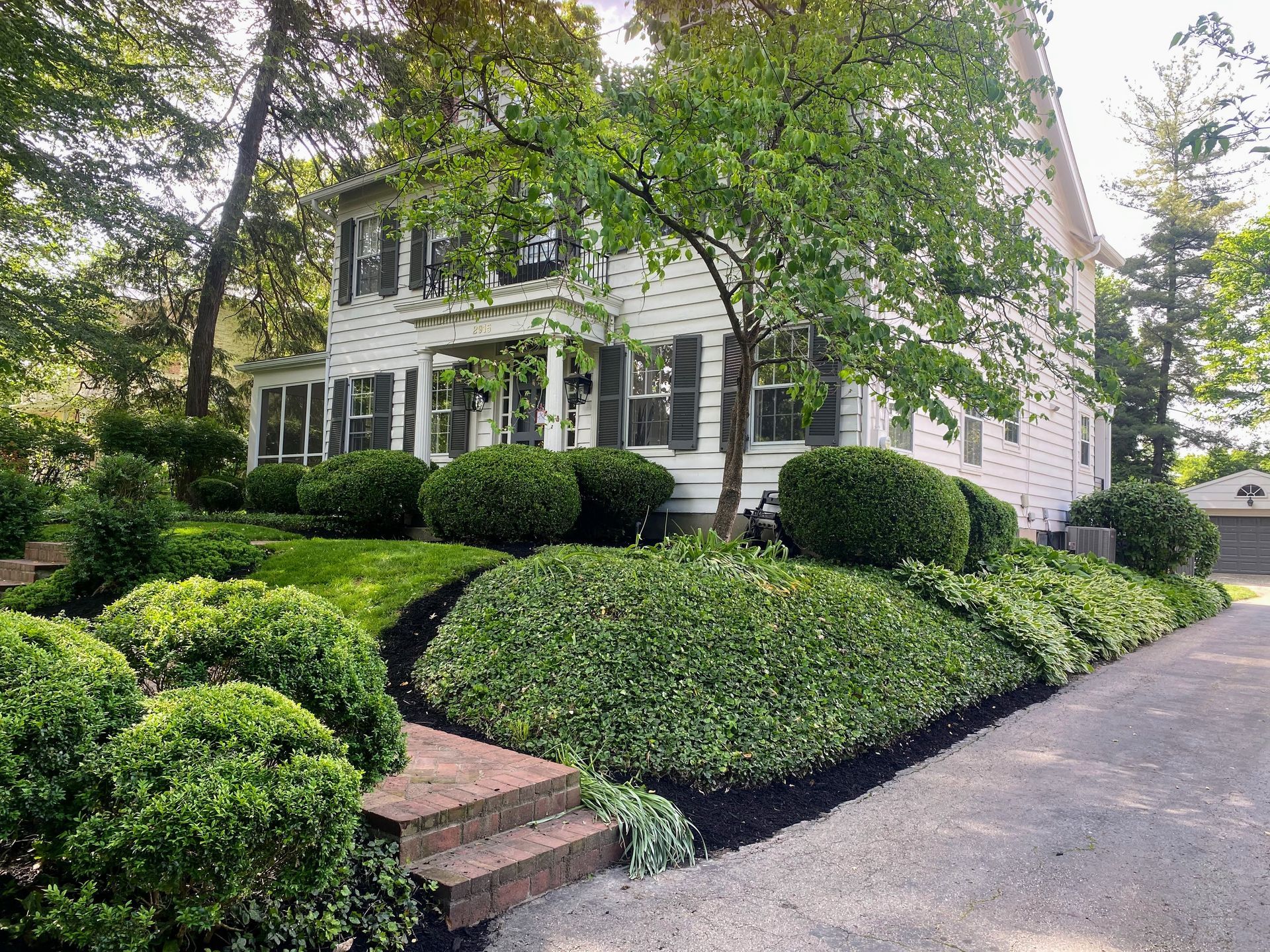 White two-story house with green landscaping, including bushes and a winding driveway. Brick steps lead up to the porch.