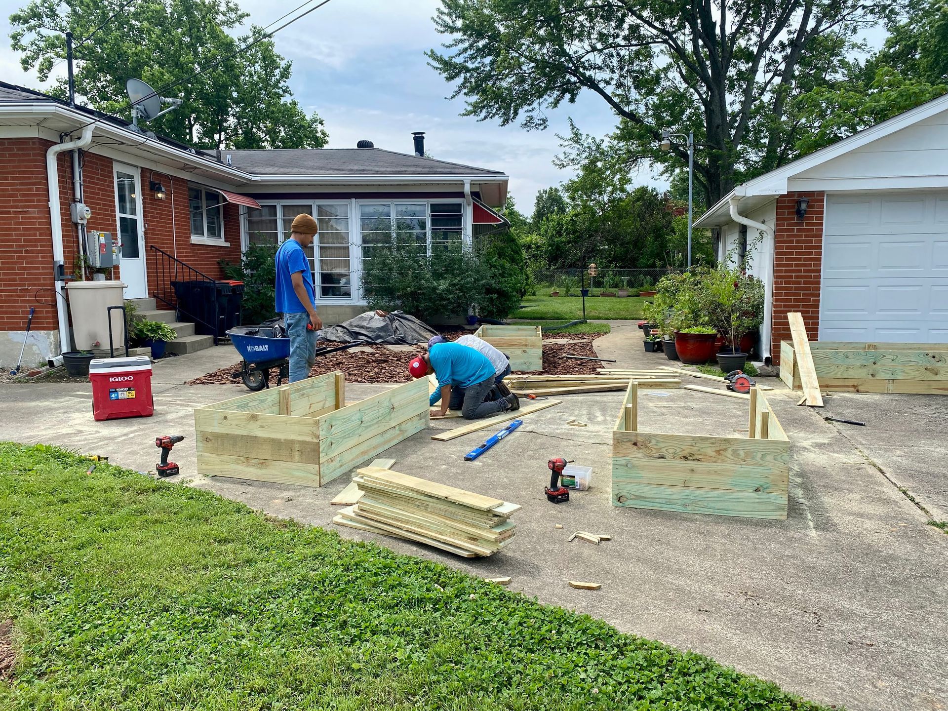 Workers building raised garden beds in a driveway beside a brick house and garage.