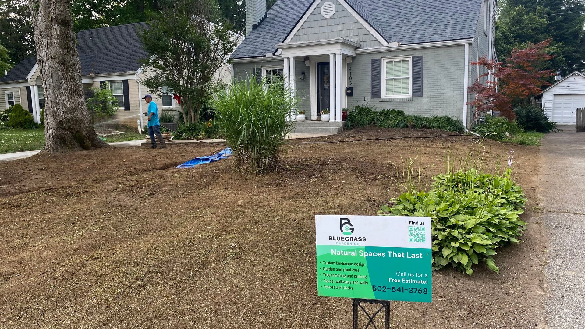 Person standing in a front yard beside a house with a lawn service sign after landscaping work